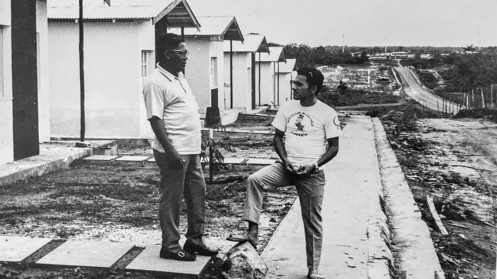 Fotografia em preto e branco: Flávio Espírito Santo, de óculos e camisa clara, conversa com um jovem diante de uma fileira de casas recém-construídas no conjunto Dom Pedro I.